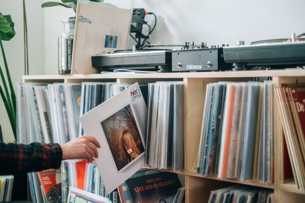 A collection of vynil records in a wooden rack with two shelves. On top of the shelf there are two record players. On hand comes from the left and selects one record titled "her".