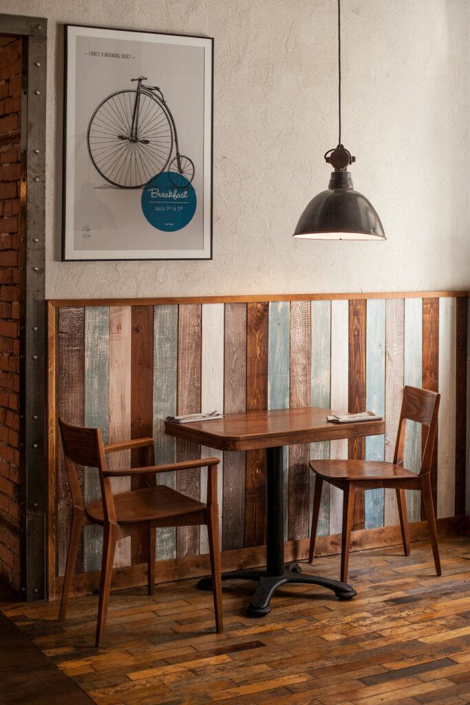 A table and two wooden chairs, possibly in a coffee shop. There's napkins and cutlery for two. The floor is wooden and the wall is half-panelled in wood too, in two tones of brown, blue-grey and white. A poster with a penny farthing image shows the breakfast times. A lamp hungs from the ceiling.